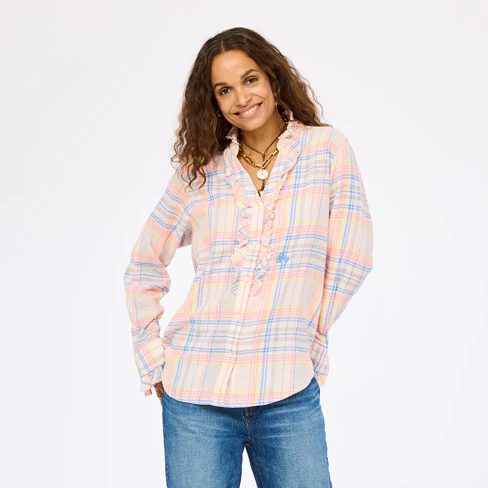 A woman with wavy brown hair smiles, wearing the Kerri Rosenthal Molly Shirt with ruffle front details and blue jeans, standing against a plain white background.