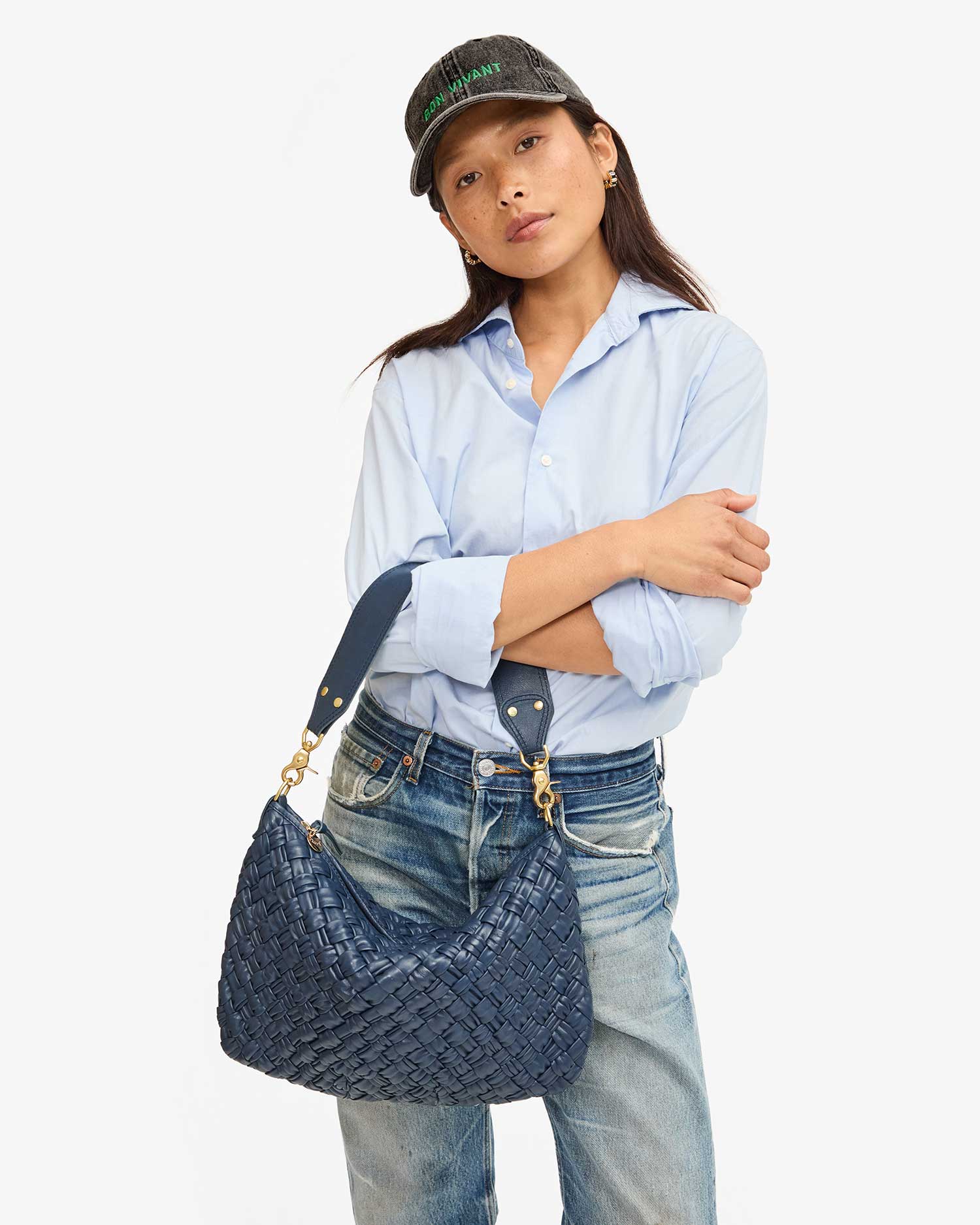 A person dressed in a light blue shirt, blue jeans, and a dark cap stands with arms crossed, holding the Moyen Messenger by Clare Vivier. The background is plain white.