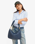 A person dressed in a light blue shirt, blue jeans, and a dark cap stands with arms crossed, holding the Moyen Messenger by Clare Vivier. The background is plain white.