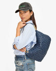 A person in a light blue shirt and jeans poses with the Clare Vivier Moyen Messenger, a chic nappa woven leather bag, slung over their shoulder. They wear a black baseball cap with green lettering. The stylish Moyen Messenger features a zip-top crescent design on a simple crossbody strap, set against the clean white background for contrast.