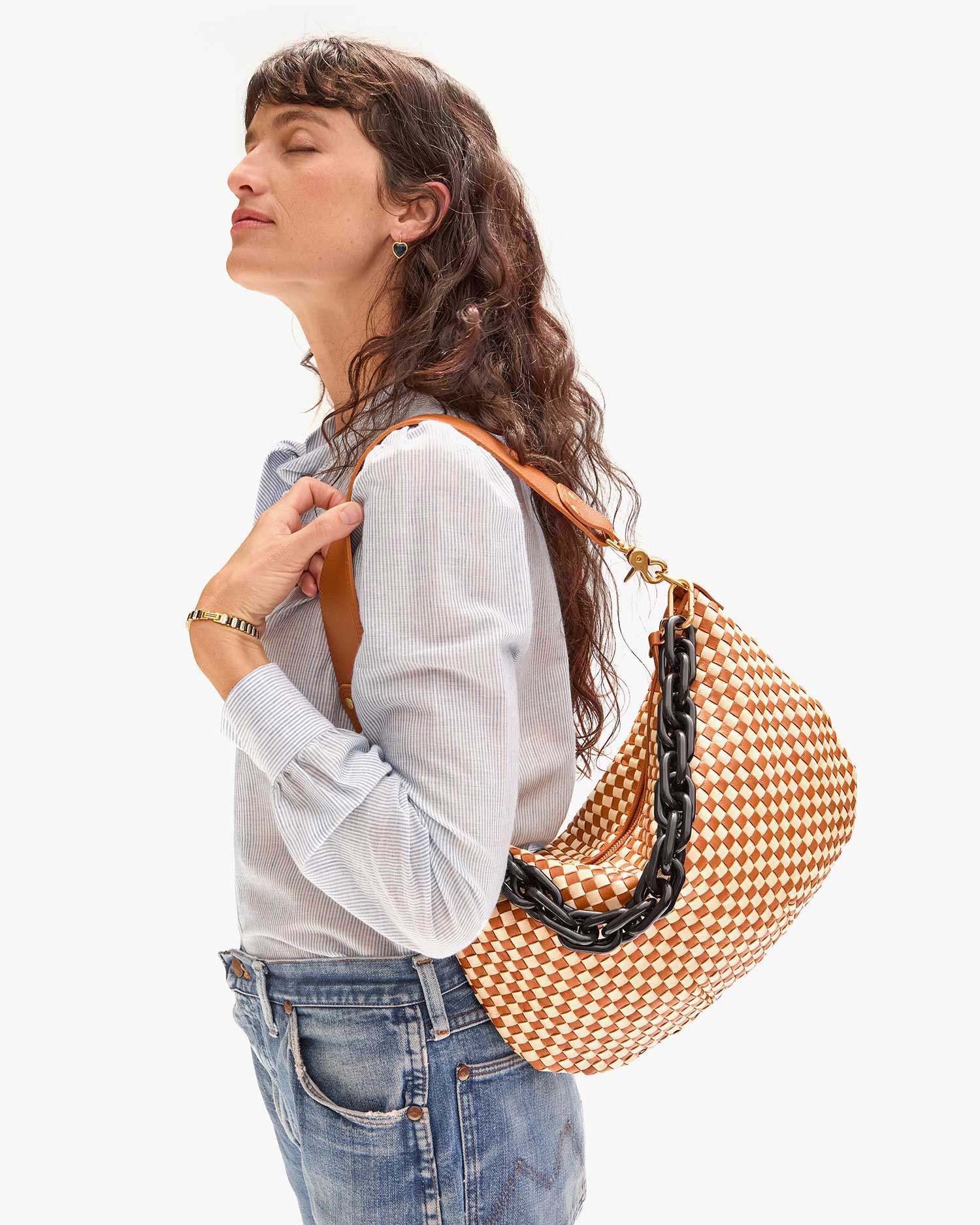 A woman with long, wavy hair stands with her eyes closed, holding the Clare Vivier Moyen Messenger bag—a brown and white checkered design with a chunky crossbody strap. She wears a light blue striped shirt and blue jeans against a plain white background.