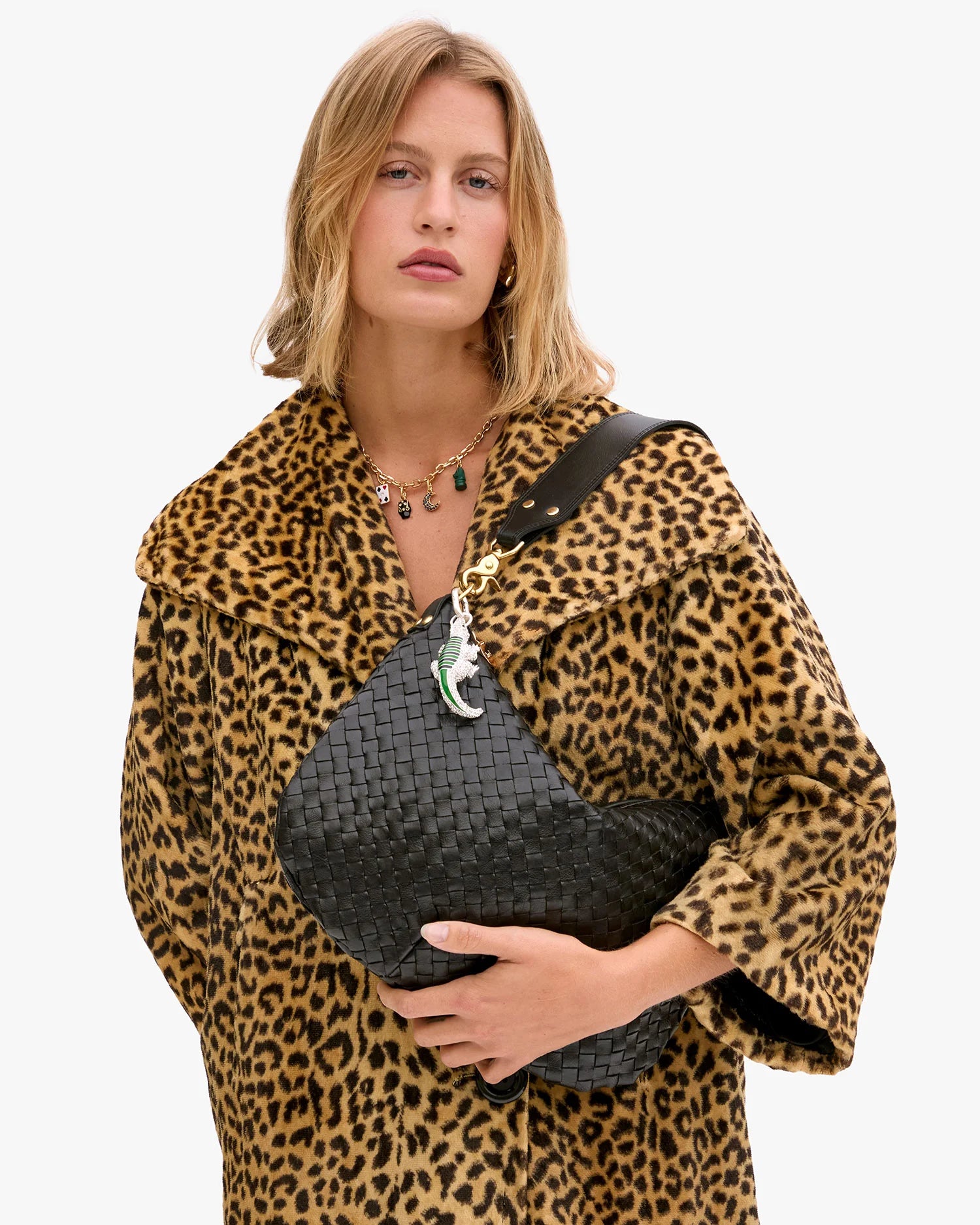 A woman in a leopard print coat holds the Clare Vivier Moyen Messenger S25, a large handwoven leather crossbody bag. She has shoulder-length blonde hair, wears a multi-pendant necklace, and stands against a plain white background.