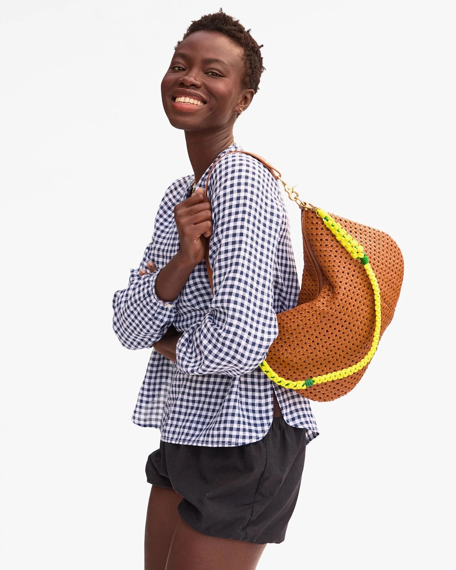 A smiling person in a blue and white checkered blouse and black shorts carries the Clare Vivier Shoulder Strap S25, a brown perforated sporty bag accessory with a bright yellow sailcord strap, over their shoulder against a plain background.