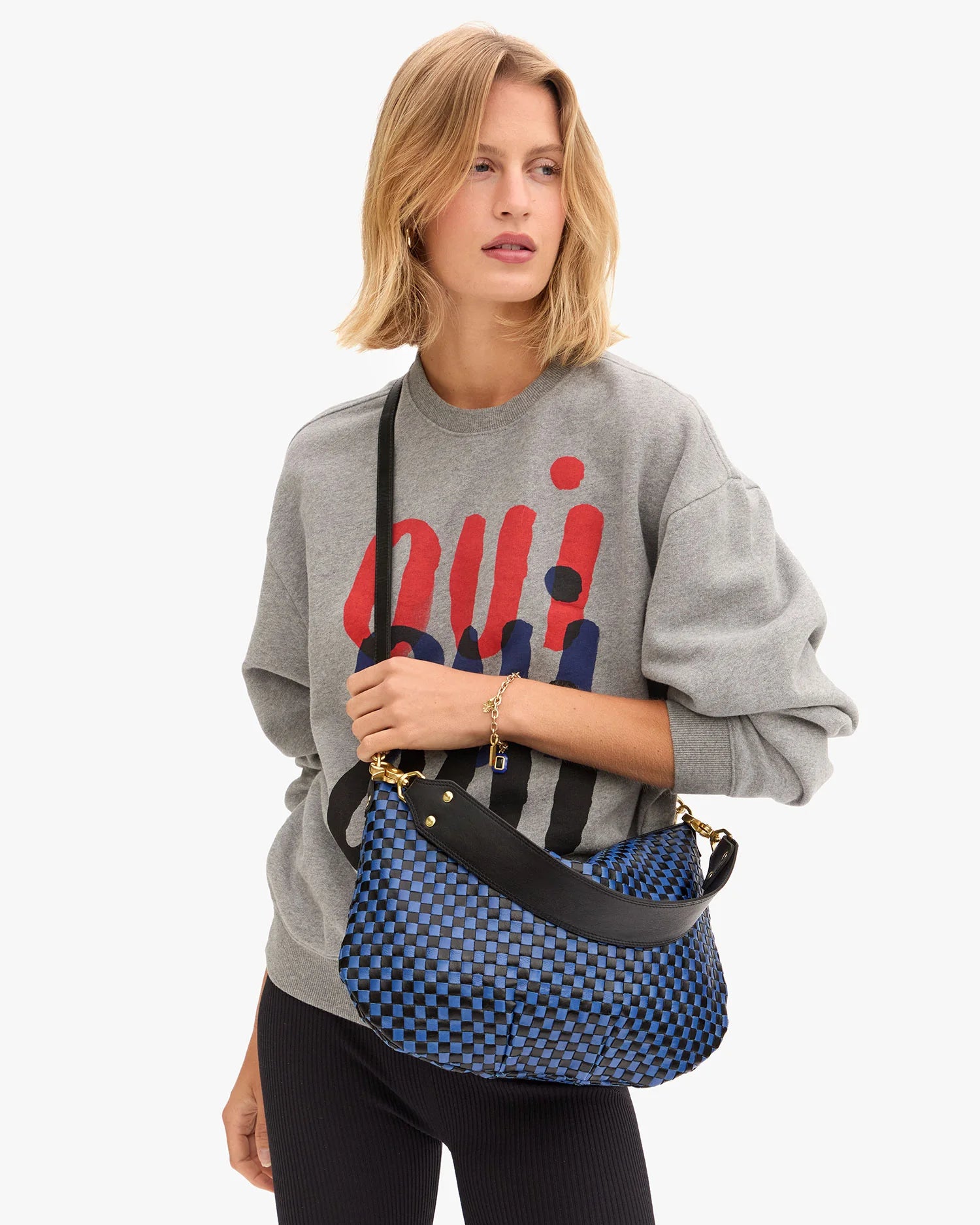 A person with shoulder-length blonde hair wears a gray sweatshirt with a red and blue abstract design, black pants, and carries the Clare Vivier Moyen Messenger S25, a blue and black checkered crescent bag with a black crossbody strap.