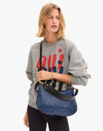 A person with shoulder-length blonde hair wears a gray sweatshirt with a red and blue abstract design, black pants, and carries the Clare Vivier Moyen Messenger S25, a blue and black checkered crescent bag with a black crossbody strap.