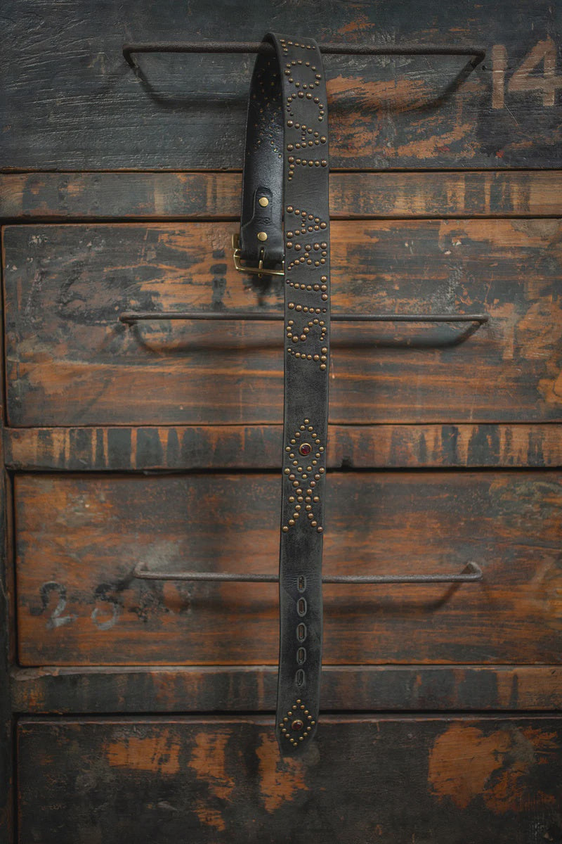 The Local Artist Belt by Made Worn, featuring metal studs and decorative patterns, hangs from the handle of a rustic wooden cabinet with distressed, dark brown drawers.