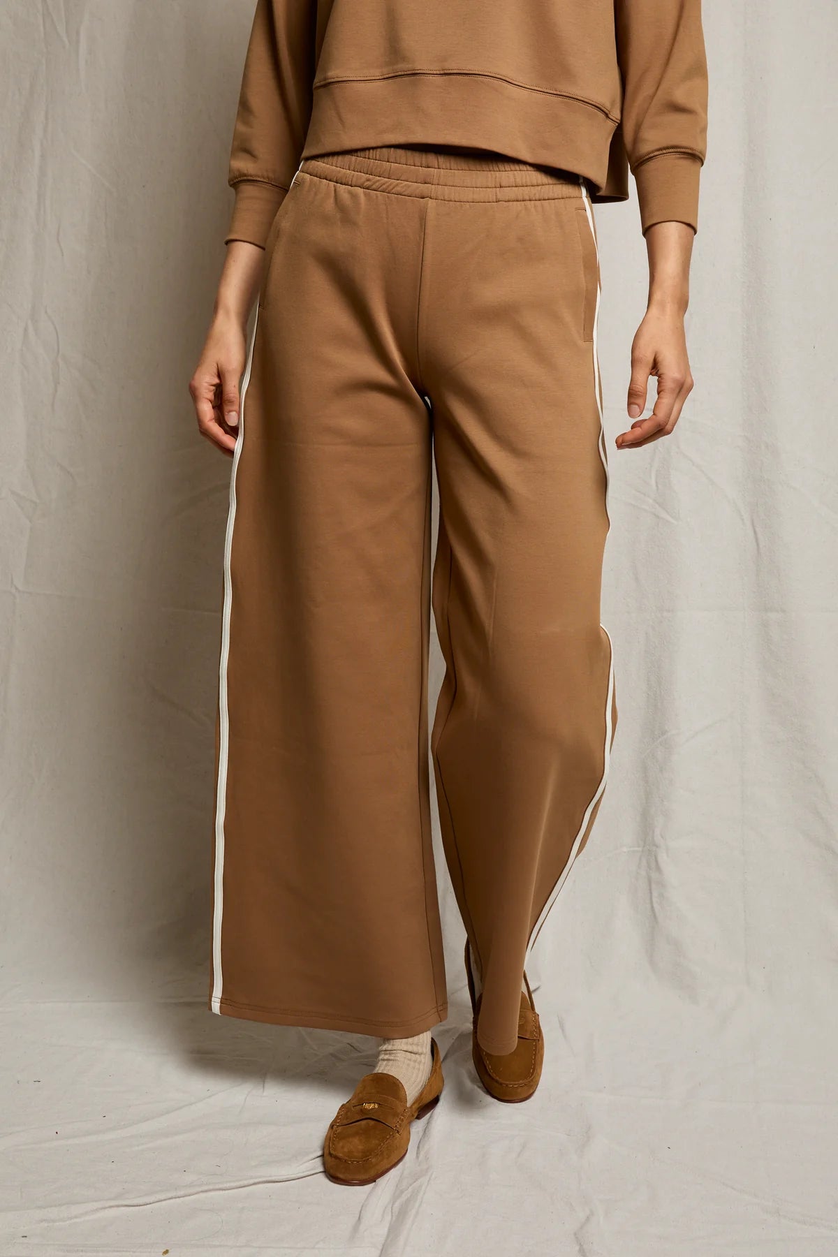 A person wears the Perfectwhitetee Mikos Travel Wideleg Pant in brown with white side stripes, paired with a matching top, beige socks, and brown loafers, standing against a neutral fabric backdrop.