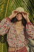 A smiling person stands in sunlight before large palm leaves, wearing the colorful Amelia Top by natalie martin and lifting a woven hat above their eyes.
