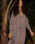 A woman with long curly hair stands among tropical plants, wearing the natalie martin MB Dress—a loose, patterned V-neck midi dress—with a relaxed expression.