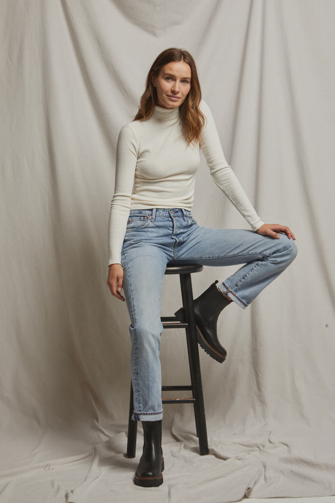 A person with long hair, dressed in the Noelle Sweater Rib LS Turtleneck by Perfectwhitetee and blue jeans, sits casually on a black stool against a simple grey draped backdrop. They wear black ankle boots and maintain a relaxed expression.