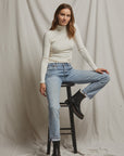 A person with long hair, dressed in the Noelle Sweater Rib LS Turtleneck by Perfectwhitetee and blue jeans, sits casually on a black stool against a simple grey draped backdrop. They wear black ankle boots and maintain a relaxed expression.