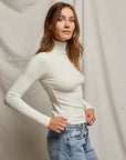 A woman with long brown hair stands in front of a plain backdrop, wearing the Noelle Sweater Rib LS Turtleneck from Perfectwhitetee paired with light blue jeans, creating a timeless wardrobe staple. She looks forward with a neutral expression.