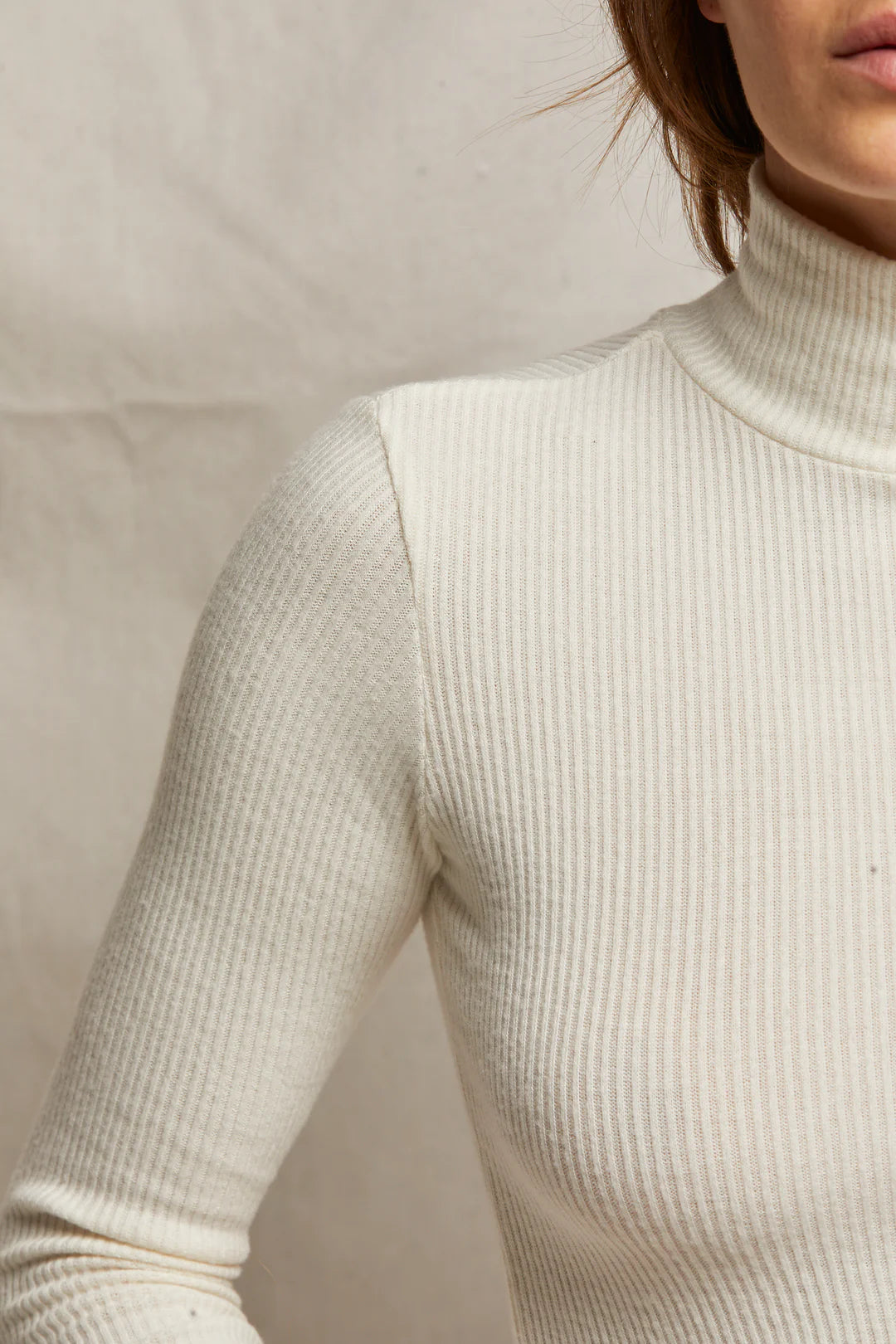 A close-up highlights the timeless silhouette of the Noelle Sweater Rib LS Turtleneck from Perfectwhitetee, as worn by a person in a cream color. The photo focuses on their upper torso and arm, with a softly blurred background. The lighting enhances this wardrobe staple's texture and elegance.