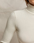 A close-up highlights the timeless silhouette of the Noelle Sweater Rib LS Turtleneck from Perfectwhitetee, as worn by a person in a cream color. The photo focuses on their upper torso and arm, with a softly blurred background. The lighting enhances this wardrobe staple's texture and elegance.