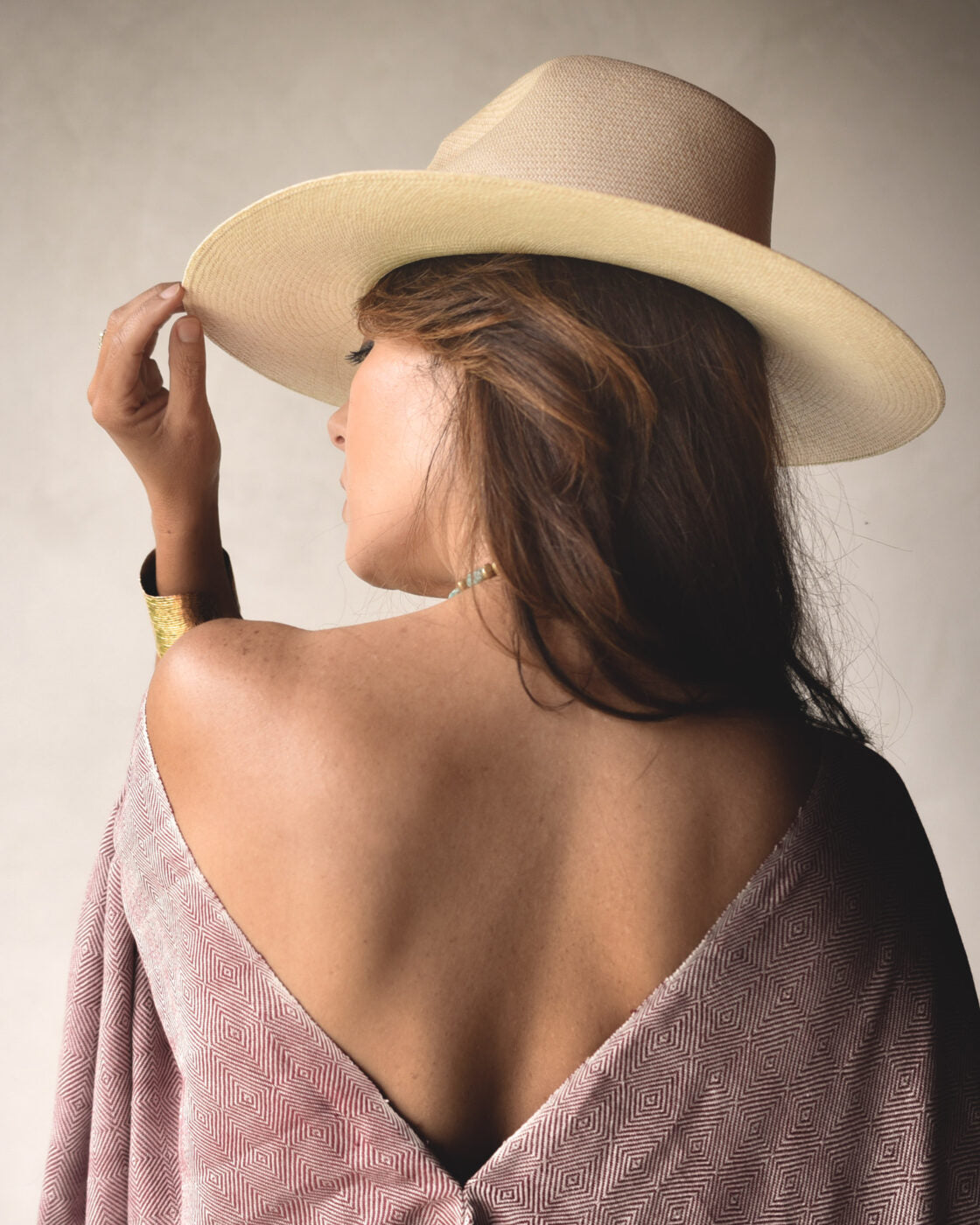 A woman with long hair is seen from behind, adjusting her Ninakuru Coda Hat, a wide-brimmed design made of toquilla straw. She wears a draped shawl that exposes her shoulders against a neutral background.