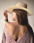 A woman with long hair is seen from behind, adjusting her Ninakuru Coda Hat, a wide-brimmed design made of toquilla straw. She wears a draped shawl that exposes her shoulders against a neutral background.