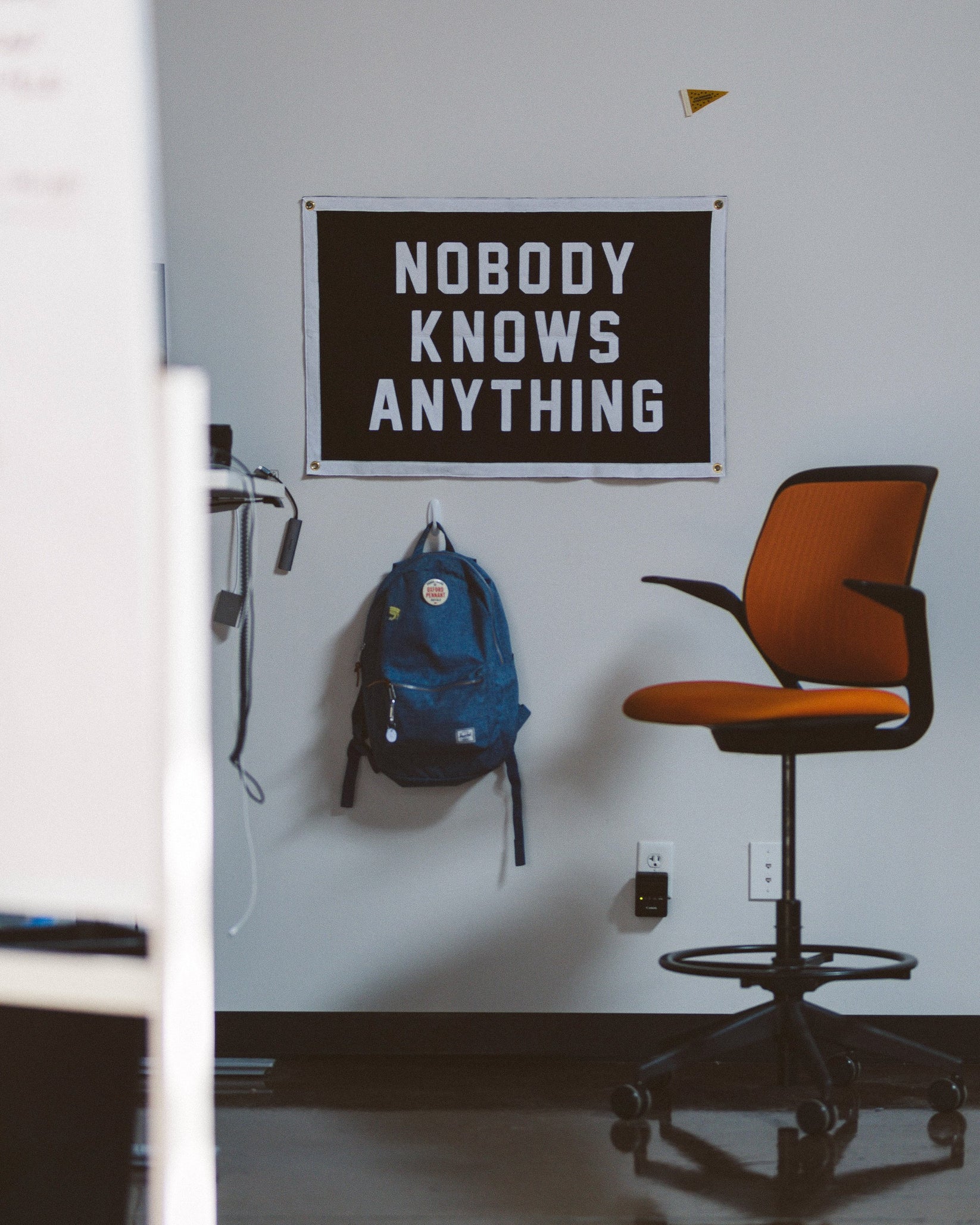 An office with a humble vibe features an orange chair, a blue backpack on the wall, and the Oxford Pennant "Nobody Knows Anything" Camp Flag in black and white.