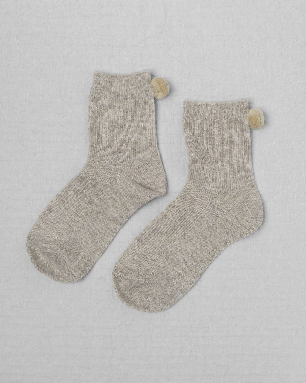 Unified’s Winter Pom Pom Socks feature a light gray crew design with small beige pompom accents at the back, displayed flat to highlight their subtle natural hues.