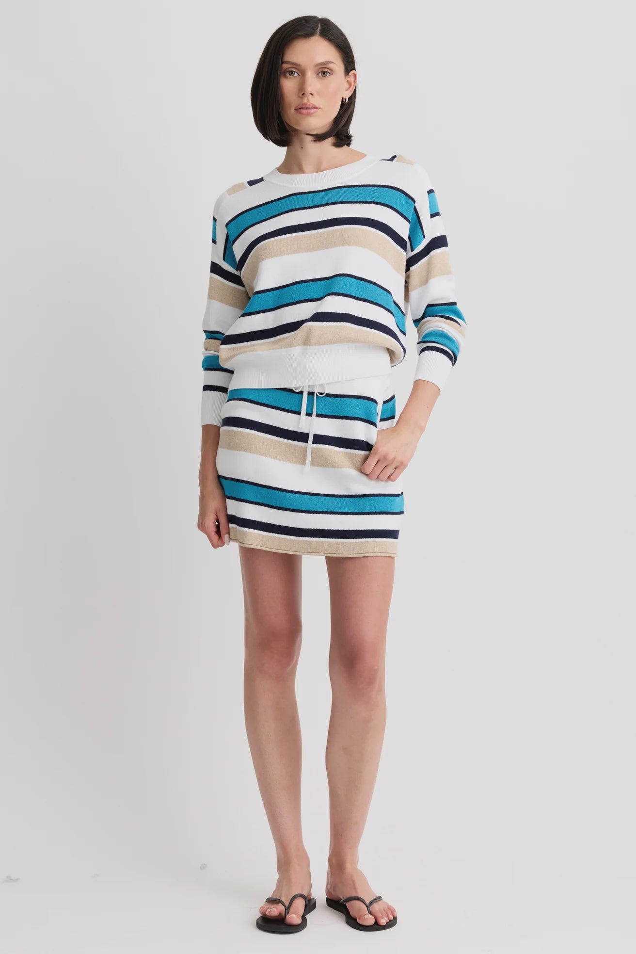 A woman stands against a plain background wearing the One Grey Day Warwick Mini Skirt and matching knit sweater in modern blue, black, white, and beige stripes, paired with black sandals.