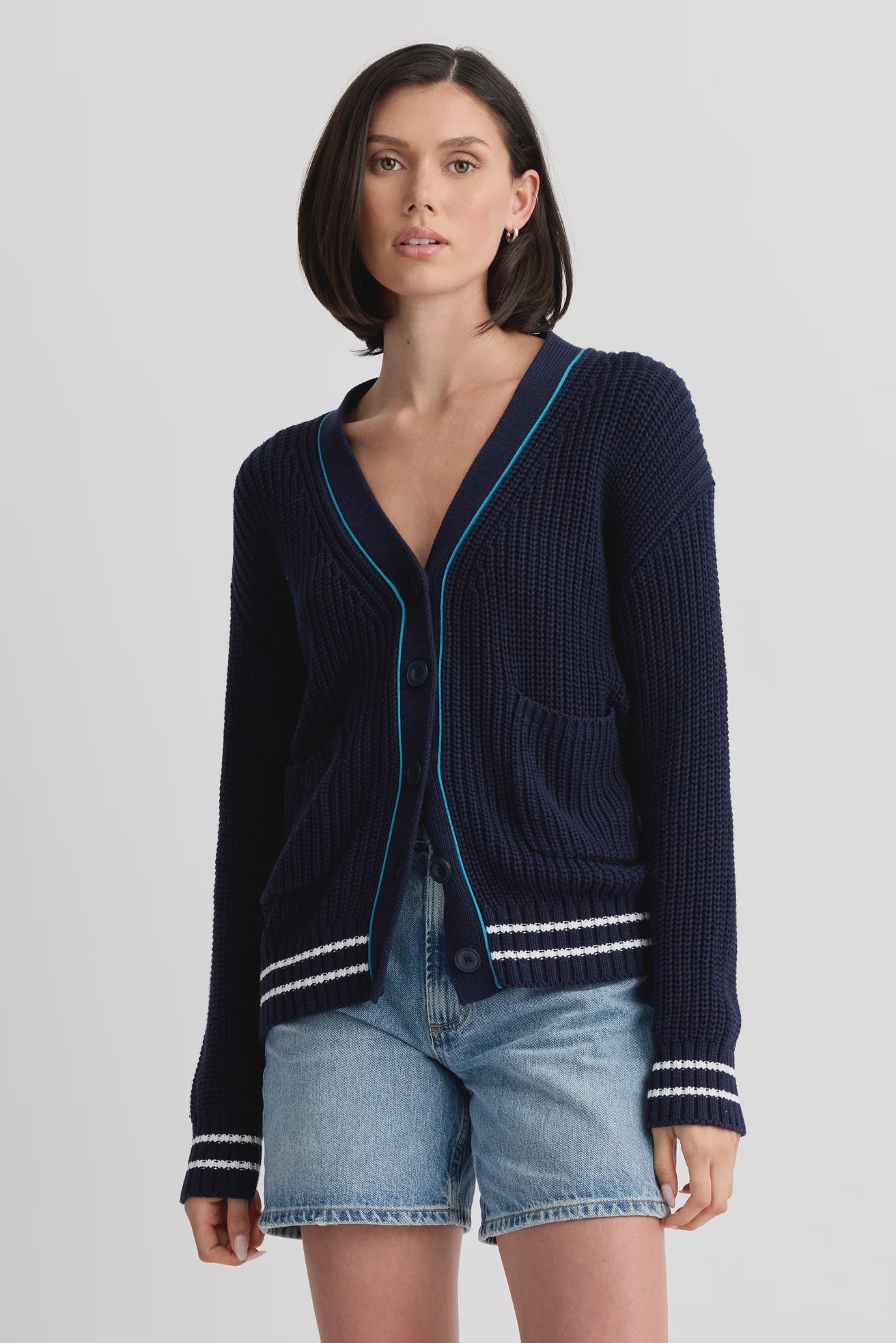 A woman with a bob haircut wears the Chelsea Cardigan by One Grey Day, featuring striped tipping and a half-cardigan stitch, over denim shorts against a plain light background.