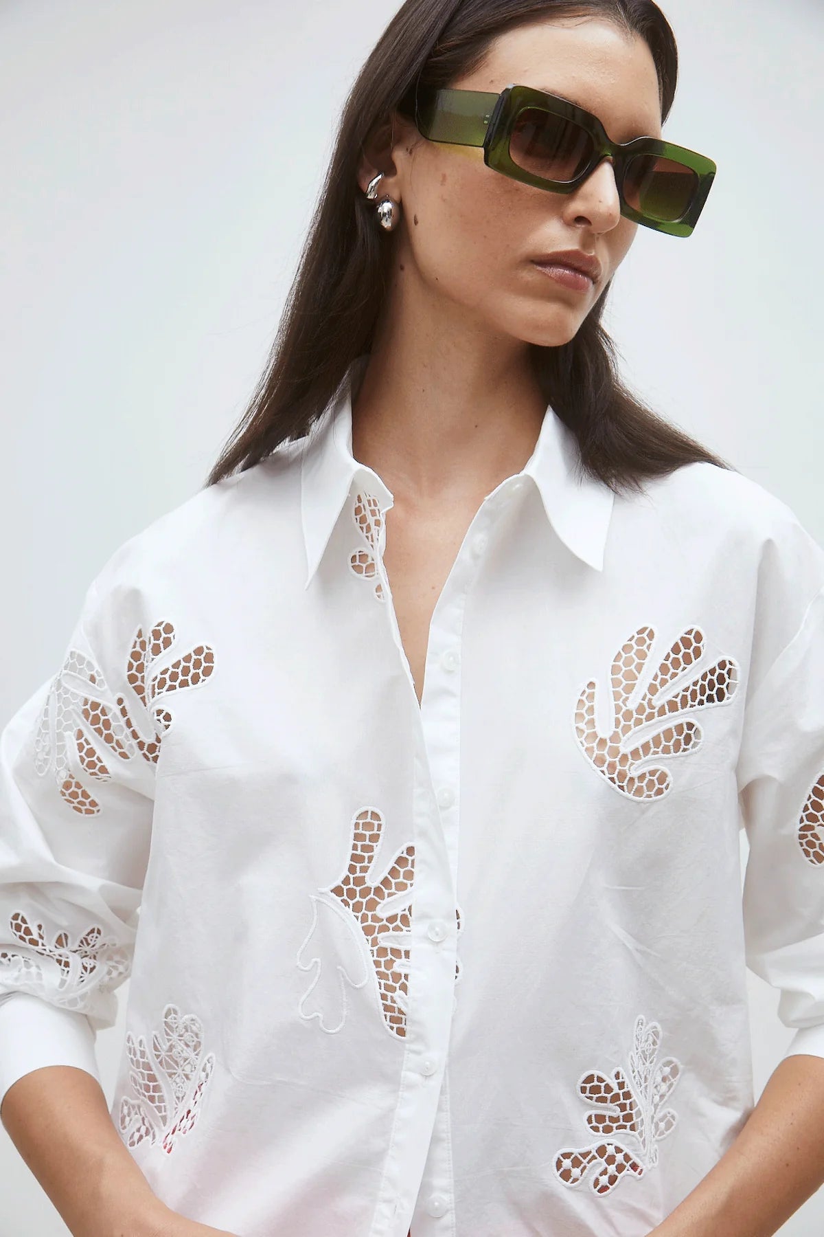 A woman wears the Loula Blouse by SUNCOO—a white button-up with lace cutouts made from 100% organic cotton—paired with oversized square sunglasses and pearl earrings, standing against a plain background.