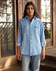 A woman with dark hair stands before a wooden building, wearing Hey Gang’s The Work Shirt—a boxy fit, light blue denim button-up with rolled sleeves—paired with ripped jeans. She looks directly at the camera.