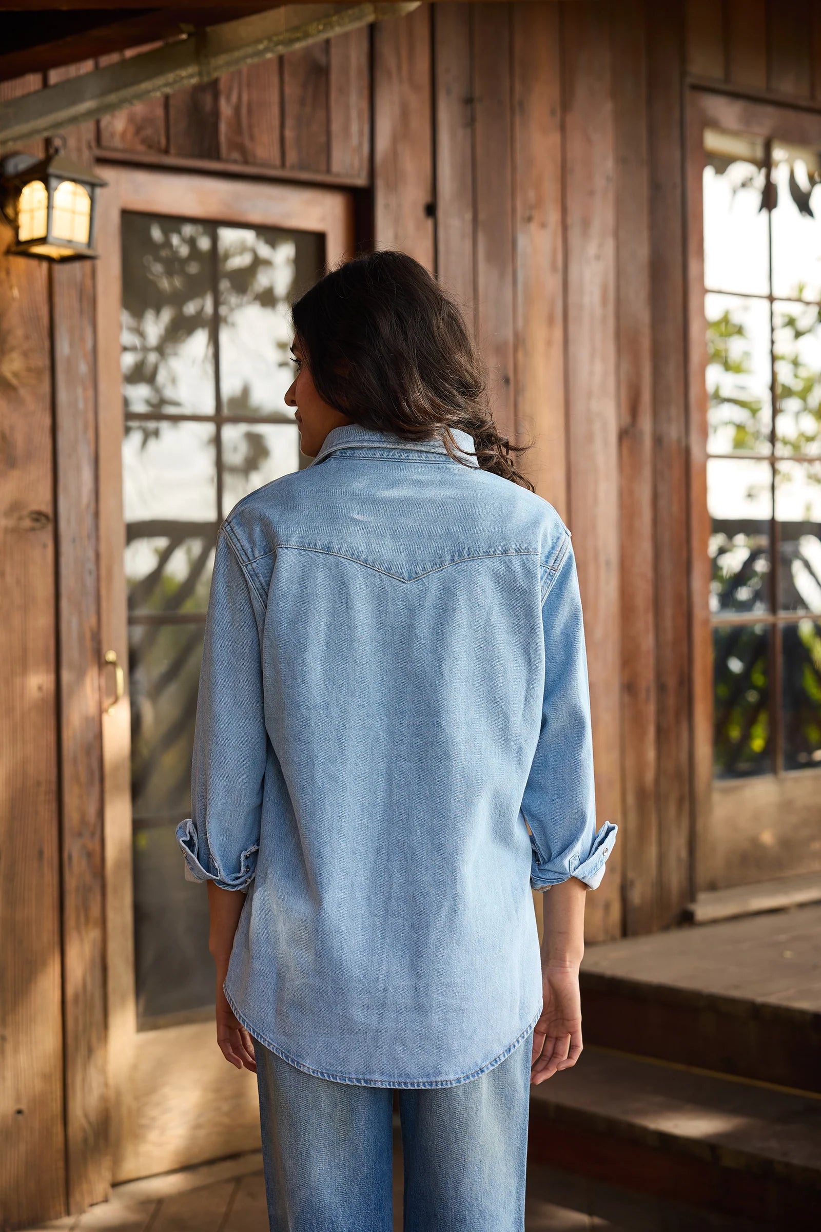 A person with long dark hair, wearing Hey Gang’s The Work Shirt in a boxy fit and jeans, stands with their back to the camera in front of a rustic wooden cabin with glass doors.