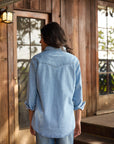 A person with long dark hair, wearing Hey Gang’s The Work Shirt in a boxy fit and jeans, stands with their back to the camera in front of a rustic wooden cabin with glass doors.
