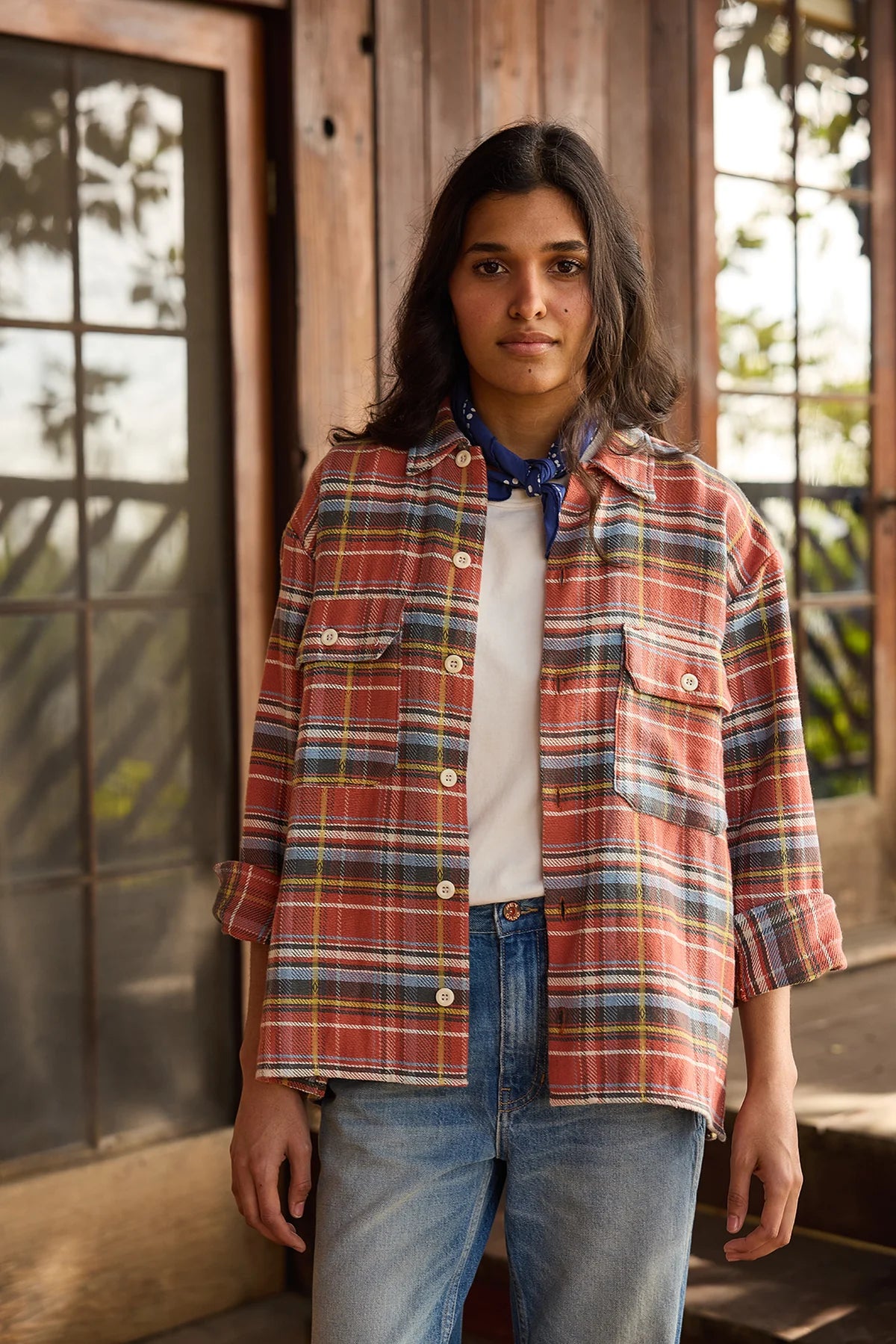 A woman with long dark hair stands outdoors by wooden doors, wearing a red plaid shirt over a white tee, blue jeans, and The HG Bandana by Hey Gang around her neck. She has a neutral expression.
