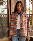 A woman with long dark hair stands outdoors by wooden doors, wearing a red plaid shirt over a white tee, blue jeans, and The HG Bandana by Hey Gang around her neck. She has a neutral expression.