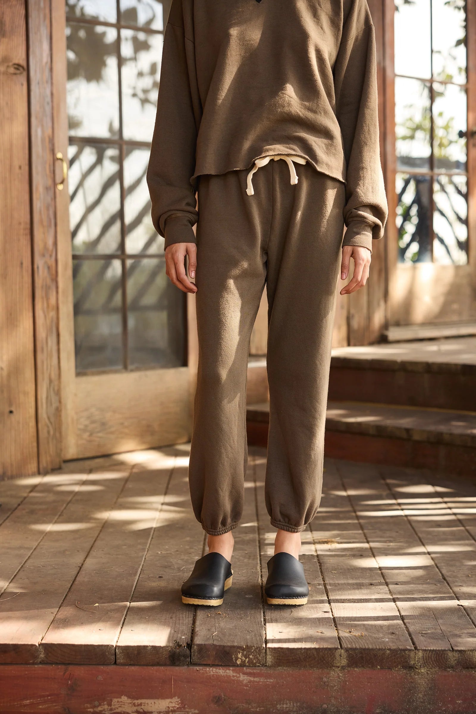 A person stands on wooden steps wearing Hey Gang&#39;s The Sweatpants in brown, paired with a matching sweatshirt and black slip-on clogs. Sunlight casts shadows on the porch and door behind them, with their upper head out of frame.