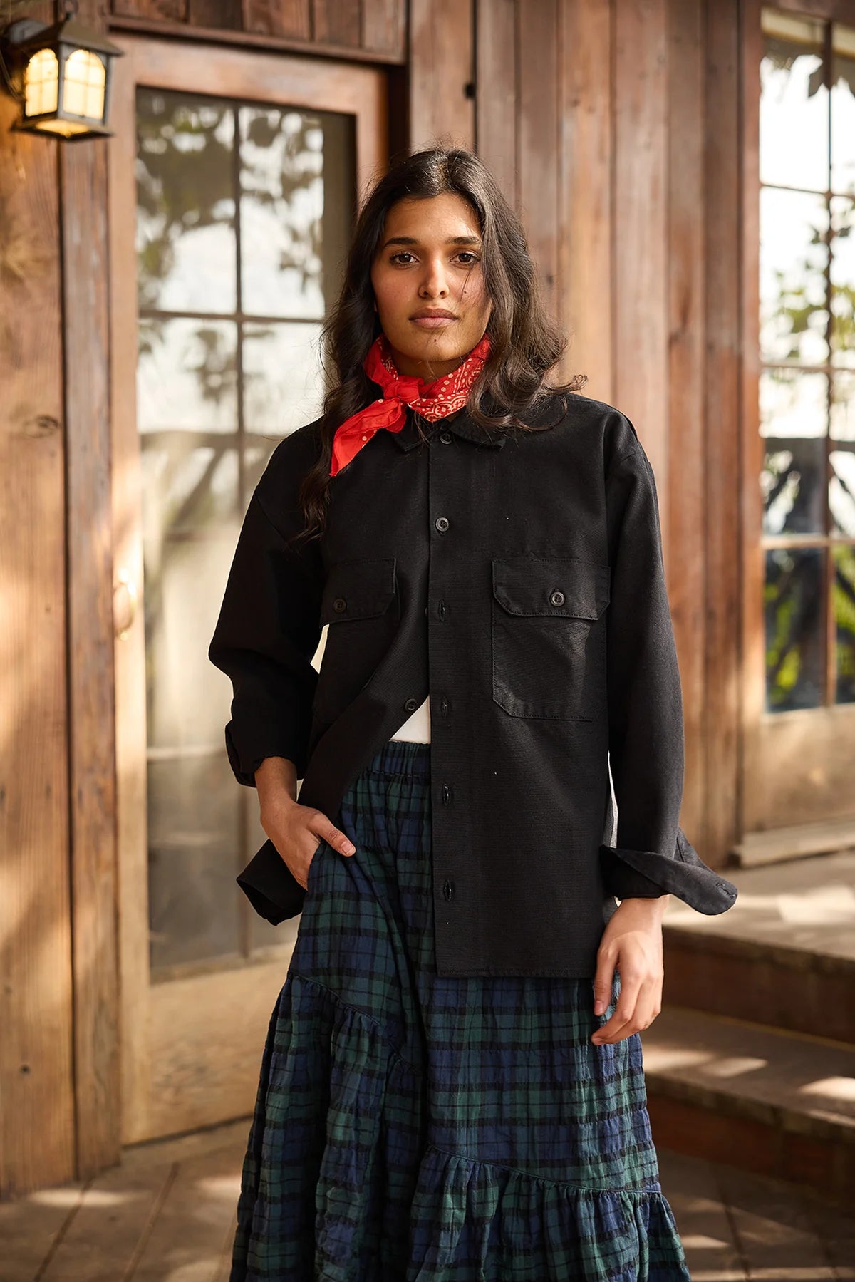 A woman with long dark hair, wearing a black button-up shirt and a blue-green plaid skirt, stands before a wooden building. Around her neck, she wears the Hey Gang HG Bandana—an everyday accessory. She gazes at the camera with a neutral expression.