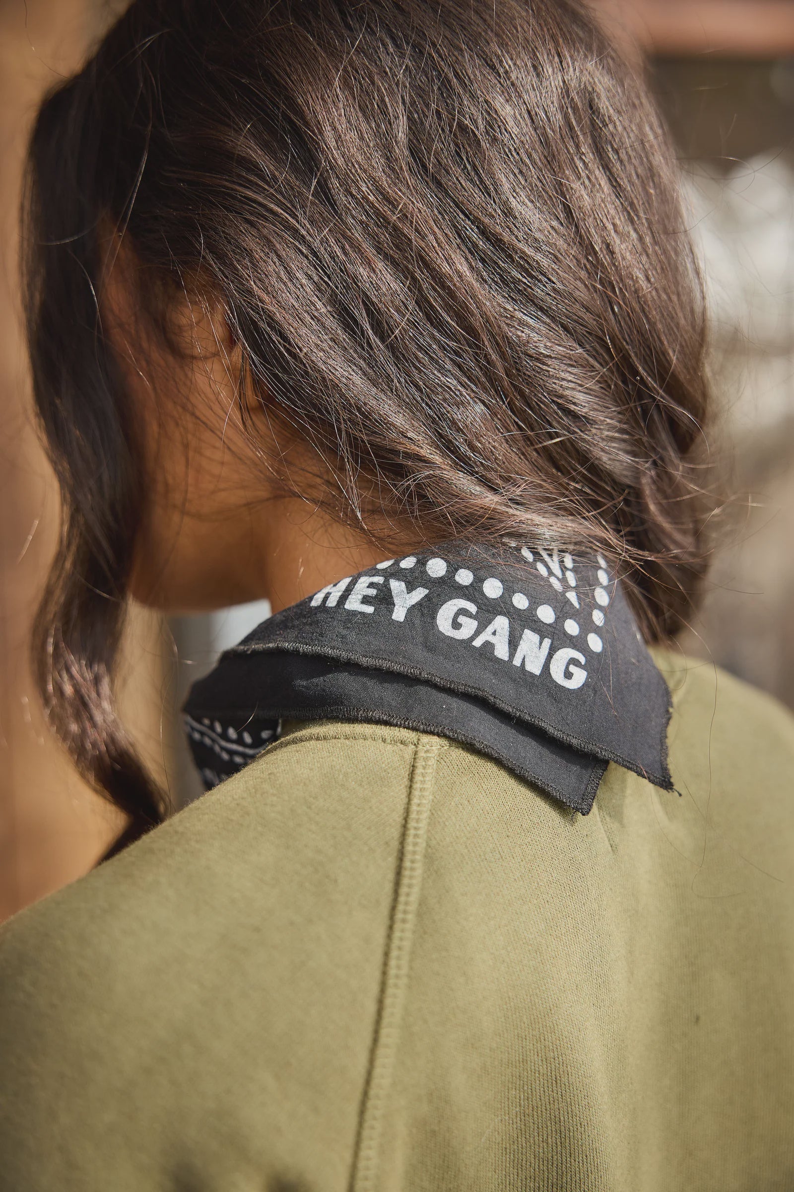 A person with long, wavy brown hair wears an olive green top and The HG Bandana by Hey Gang around their neck, which features "HEY GANG" in white letters. The image highlights the back and side of their head and shoulders.