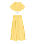 The image shows a matching yellow two-piece outfit made of 100% cotton. The top, labeled "Opihi Bubble Sleeve Crop Top" by Mikoh, is a short-sleeved, loose-fitting blouse with bubble sleeves. The bottom, labeled "DELIA," is a high-waisted, long, flowy skirt. Both pieces are displayed against a white background.