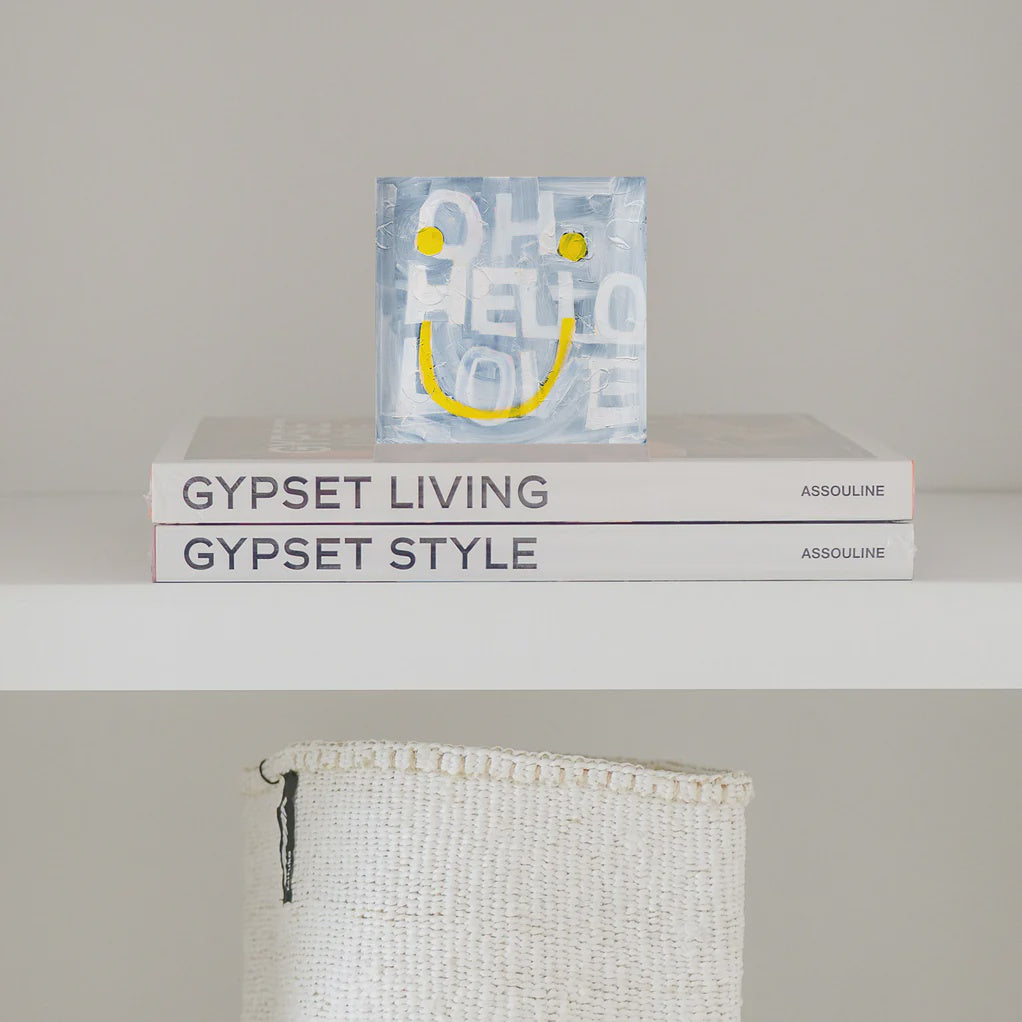 The "Oh Hello Smiley Block of Love" by Kerri Rosenthal, featuring a yellow smiley face and the text "OH HELLO LOVE," is placed on two Assouline books, "GYPSET LIVING" and "GYPSET STYLE." A sleek plexiglass artwork sits beside a white woven basket on the shelf below.