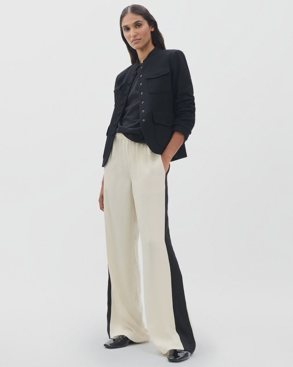 A woman stands against a white background wearing the Rag & Bone olivia stripe pant with black side panels, paired with a black button-up shirt, black jacket, and black shoes. Her long dark hair and neutral expression highlight the minimalist look.