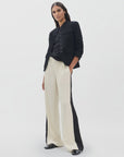 A woman stands against a white background wearing the Rag & Bone olivia stripe pant with black side panels, paired with a black button-up shirt, black jacket, and black shoes. Her long dark hair and neutral expression highlight the minimalist look.