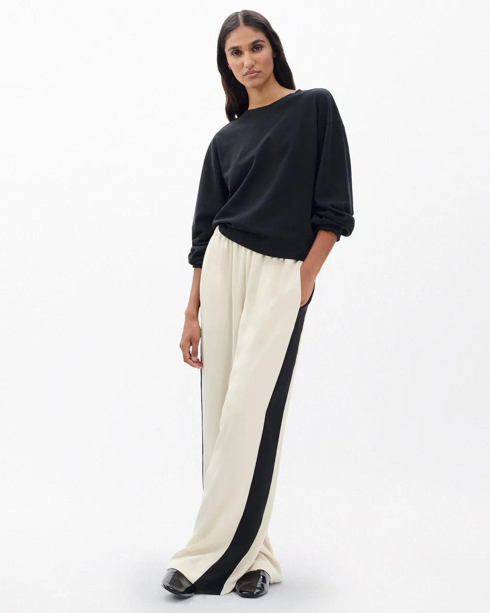 A woman poses against a plain background in the Rag & Bone olivia stripe pant—cream wide-leg pants with a black side stripe—styled with a loose black top, black shoes, and her hand in her pocket. She has long dark hair for a relaxed, sporty look.