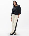 A woman poses against a plain background in the Rag & Bone olivia stripe pant—cream wide-leg pants with a black side stripe—styled with a loose black top, black shoes, and her hand in her pocket. She has long dark hair for a relaxed, sporty look.