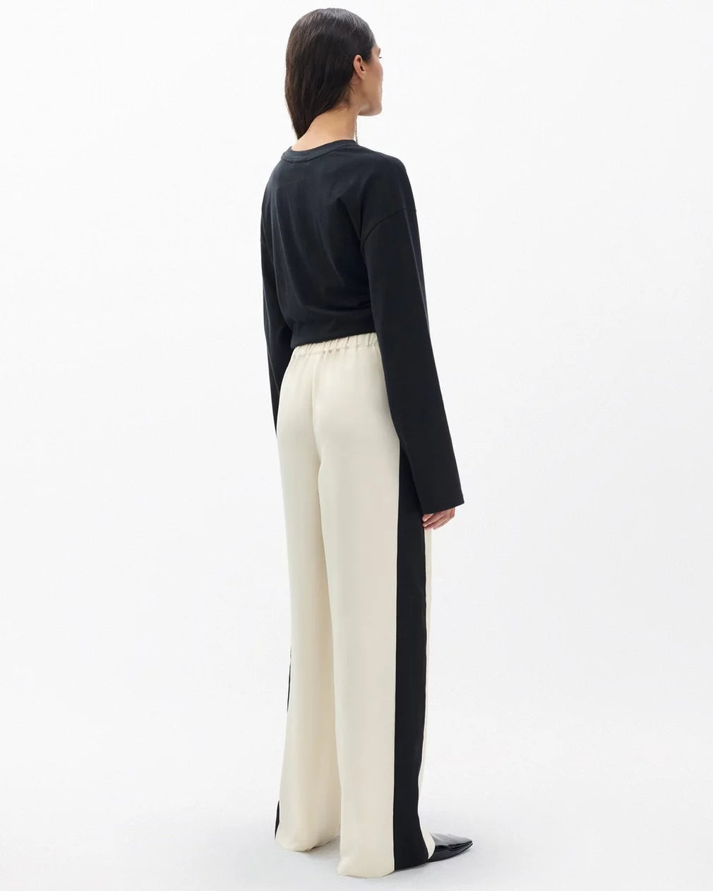 A woman faces away, wearing a black long-sleeve top and Rag & Bone's olivia stripe pant—high-waisted, wide-leg cream trousers with black side panels—against a plain white background.