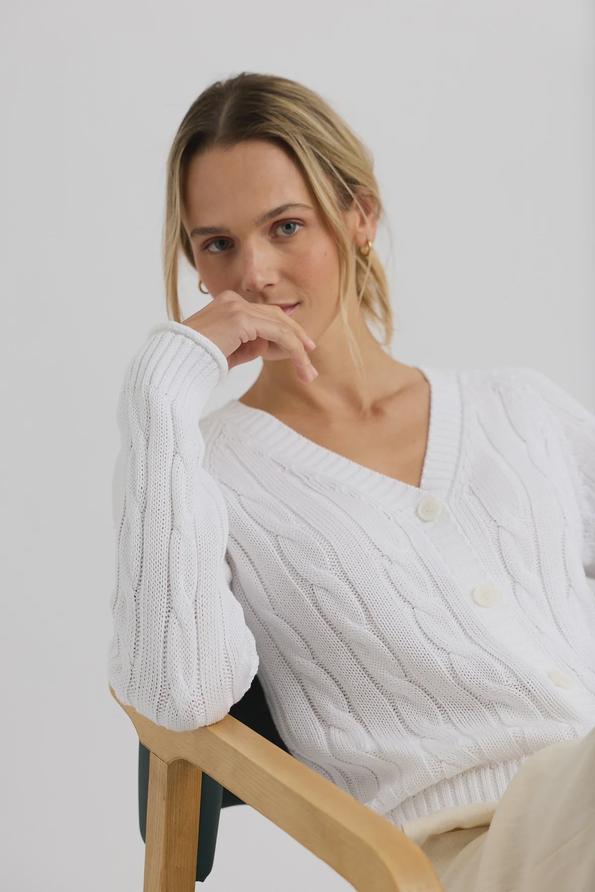 A blonde woman wearing the One Grey Day Raleigh Cable Cardigan and light pants sits on a wooden chair, leaning her head on her hand and gazing calmly at the camera.