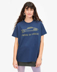 A person with long hair wears Clare Vivier's Classic Tee S24, a navy blue shirt featuring a gold fish design and the phrase "Liberez Les Sardines." They're against a neutral background, light-colored pants visible beneath the shirt, seemingly ready to bid farewell to ordinary fashion.