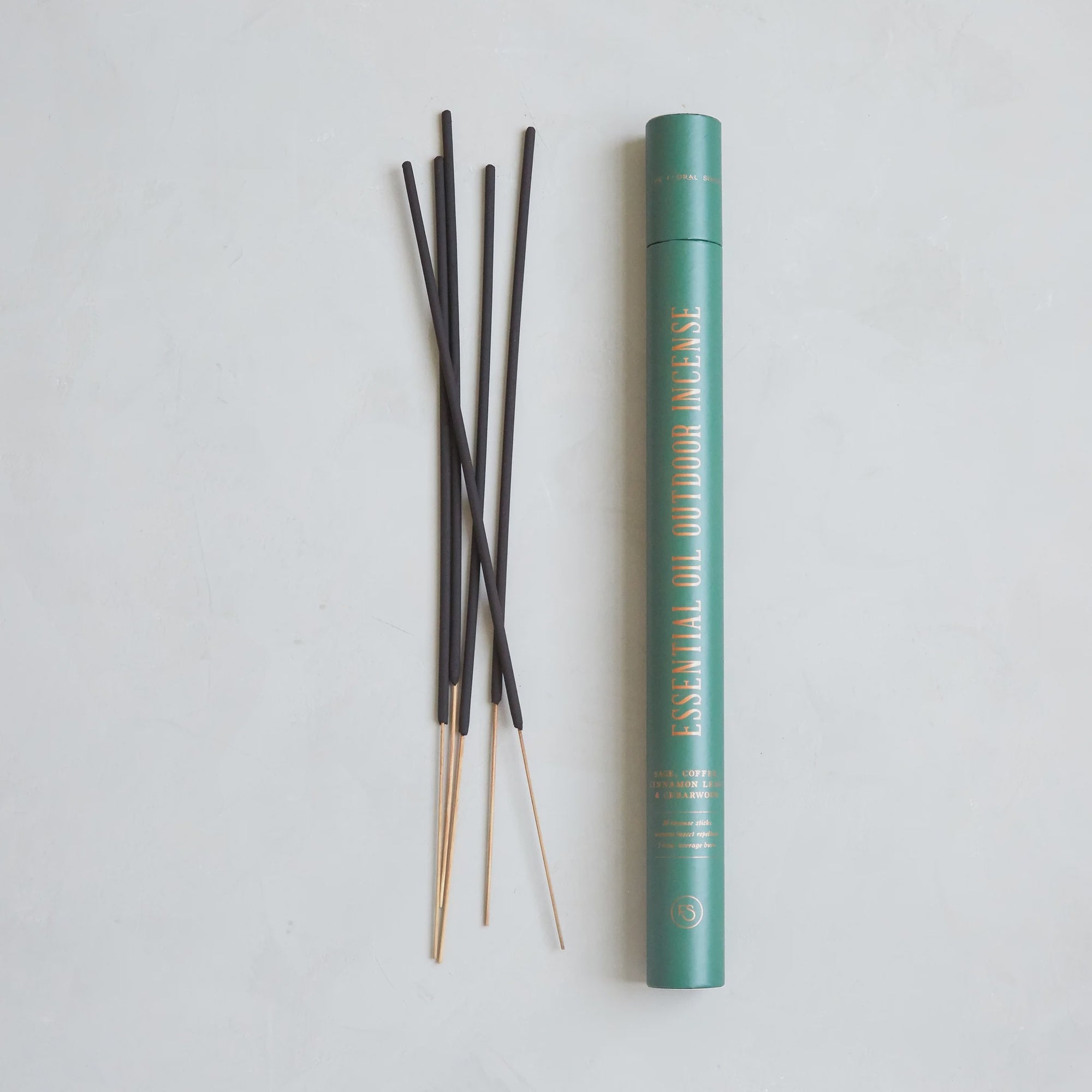 A green cylindrical container with "The Floral Society's Essential Oil Outdoor Incense," handcrafted in Connecticut, sits on a light surface. Beside it lie several black incense sticks featuring wooden ends, infused with insect-repellent essential oils.