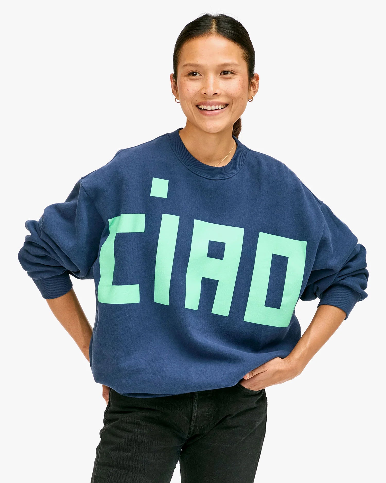 A person smiling and wearing an oversized Clare Vivier sweatshirt in dark blue, emblazoned with the word "CIAO" in large, light green letters. They confidently stand against a plain, light-colored background with their hands on their hips, exuding stylish comfort.