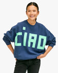 A person smiling and wearing an oversized Clare Vivier sweatshirt in dark blue, emblazoned with the word "CIAO" in large, light green letters. They confidently stand against a plain, light-colored background with their hands on their hips, exuding stylish comfort.