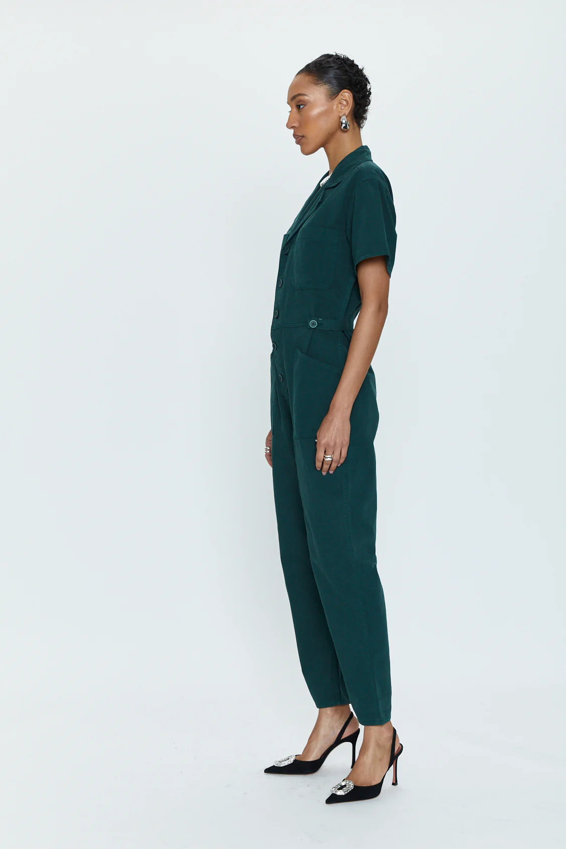 A person stands in profile dressed in Pistola's best-selling Grover Short Sleeve Field Suit, featuring a tailored fit and an emerald green shade. They are accessorized with black high-heeled shoes adorned with silver details. The deep jeweled tone of the jumpsuit contrasts beautifully against the plain white background, highlighting the attire.