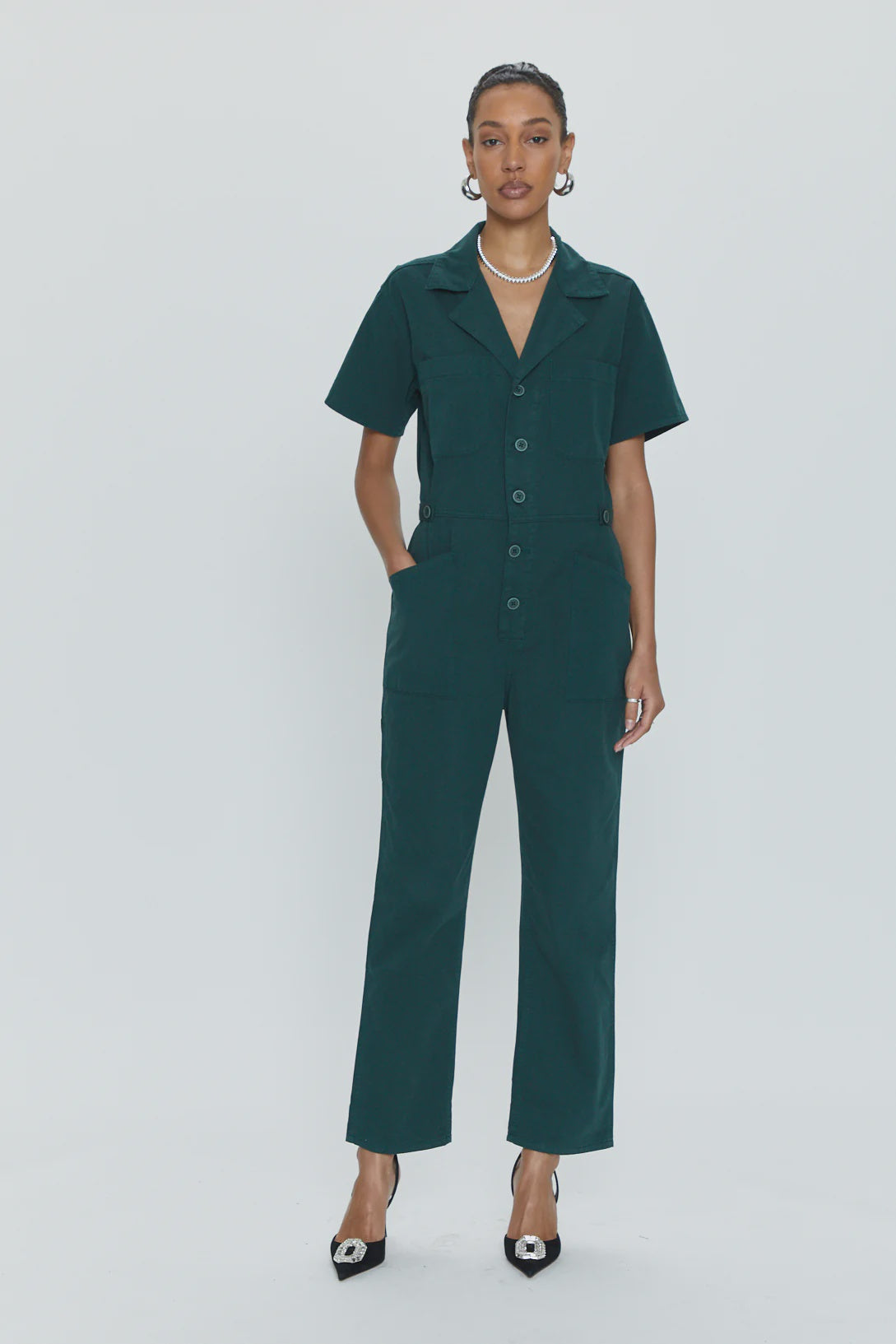 A person stands against a plain, light background wearing the Pistola Grover Short Sleeve Field Suit in dark green, characterized by its button-down front and relaxed fit. They have their hands in the pockets of the jumpsuit and accessorize with black pointed-toe heels and a pearl necklace.