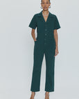 A person stands against a plain, light background wearing the Pistola Grover Short Sleeve Field Suit in dark green, characterized by its button-down front and relaxed fit. They have their hands in the pockets of the jumpsuit and accessorize with black pointed-toe heels and a pearl necklace.