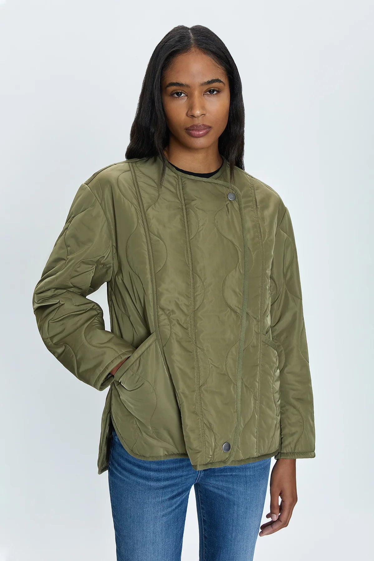 A person with long dark hair is wearing a Pistola Elva Onion Stitch Puffer Jacket in olive green, featuring onion stitch quilting, front buttons, and pockets. They have a neutral expression and are standing against a plain, light-colored background. Additionally, they are wearing blue jeans.