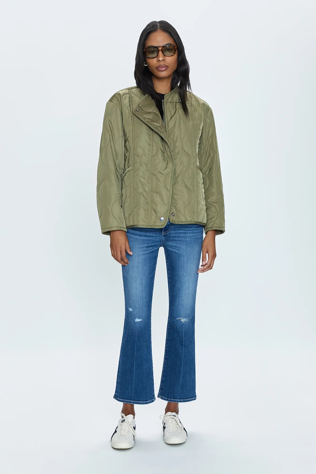 A person stands against a plain background wearing the Pistola Elva Onion Stitch Puffer Jacket in military green over a black top, paired with blue flared jeans featuring slight distressing at the knees, white sneakers, and dark sunglasses. They have long dark hair and stand with a relaxed posture.